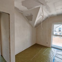 ReMalFach - Pokój w trakcie prac wykończeniowych: ściany i sufit po szpachlowaniu, zabezpieczona podłoga folią. Widoczne kable elektryczne i okno z widokiem na budowę.