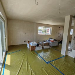 ReMalFach - Pokój w trakcie prac wykończeniowych: zabezpieczona folią podłoga, świeżo położona gładź na ścianach, okno z widokiem na zewnątrz, przygotowane materiały budowlane.