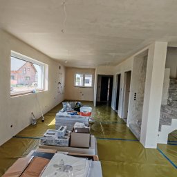 ReMalFach - Wnętrze domu w trakcie prac wykończeniowych: gładzie na ścianach i suficie, podłoga zabezpieczona folią, widoczne materiały budowlane i narzędzia. Jasne pomieszczenie z oknami.