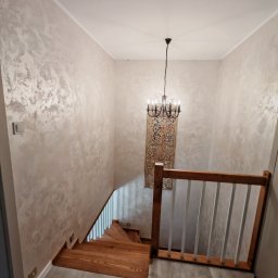 ReMalFach - Klatka schodowa z drewnianymi schodami i balustradą. Ściany pokryte dekoracyjną, jasną gładzią z subtelnym wzorem. Na ścianie ozdobny panel i żyrandol.