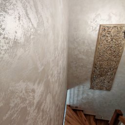 ReMalFach - Ściana z dekoracyjną, beżowo-srebrną fakturą w korytarzu, widok z góry na drewniane schody i ozdobny, drewniany panel na ścianie. Efektowna aranżacja wnętrza.