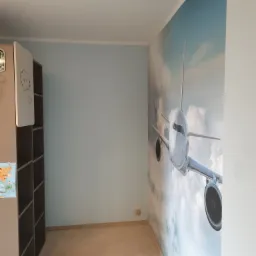 Pokój dziecięcy z fototapetą przedstawiającą samolot pasażerski w chmurach, widoczny fragment regału z rysunkami chmurek.