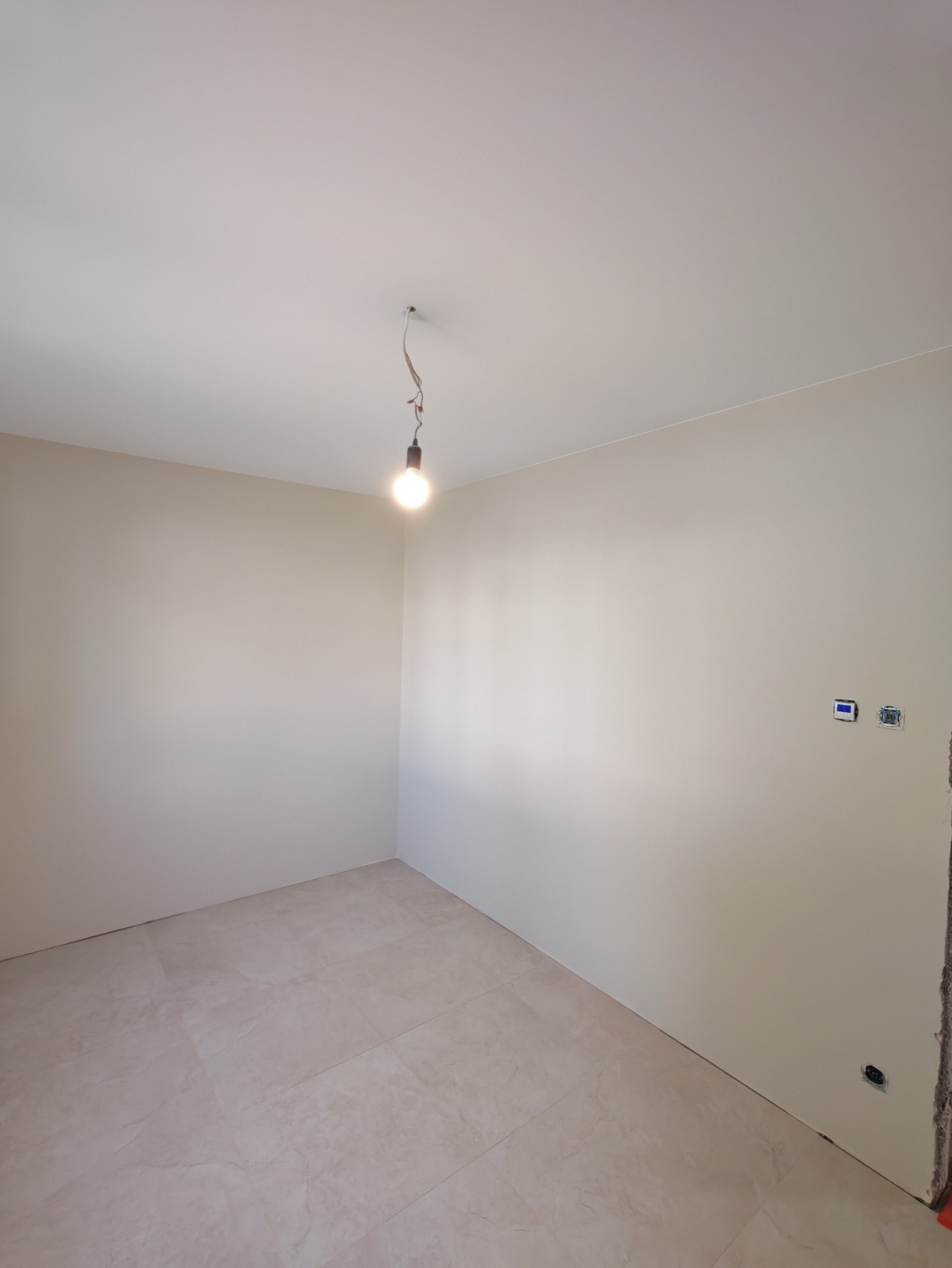 Jasne wnętrze po szpachlowaniu i malowaniu ścian. Widoczne gniazdka elektryczne, podłoga wyłożona płytkami i żarówka na kablu.