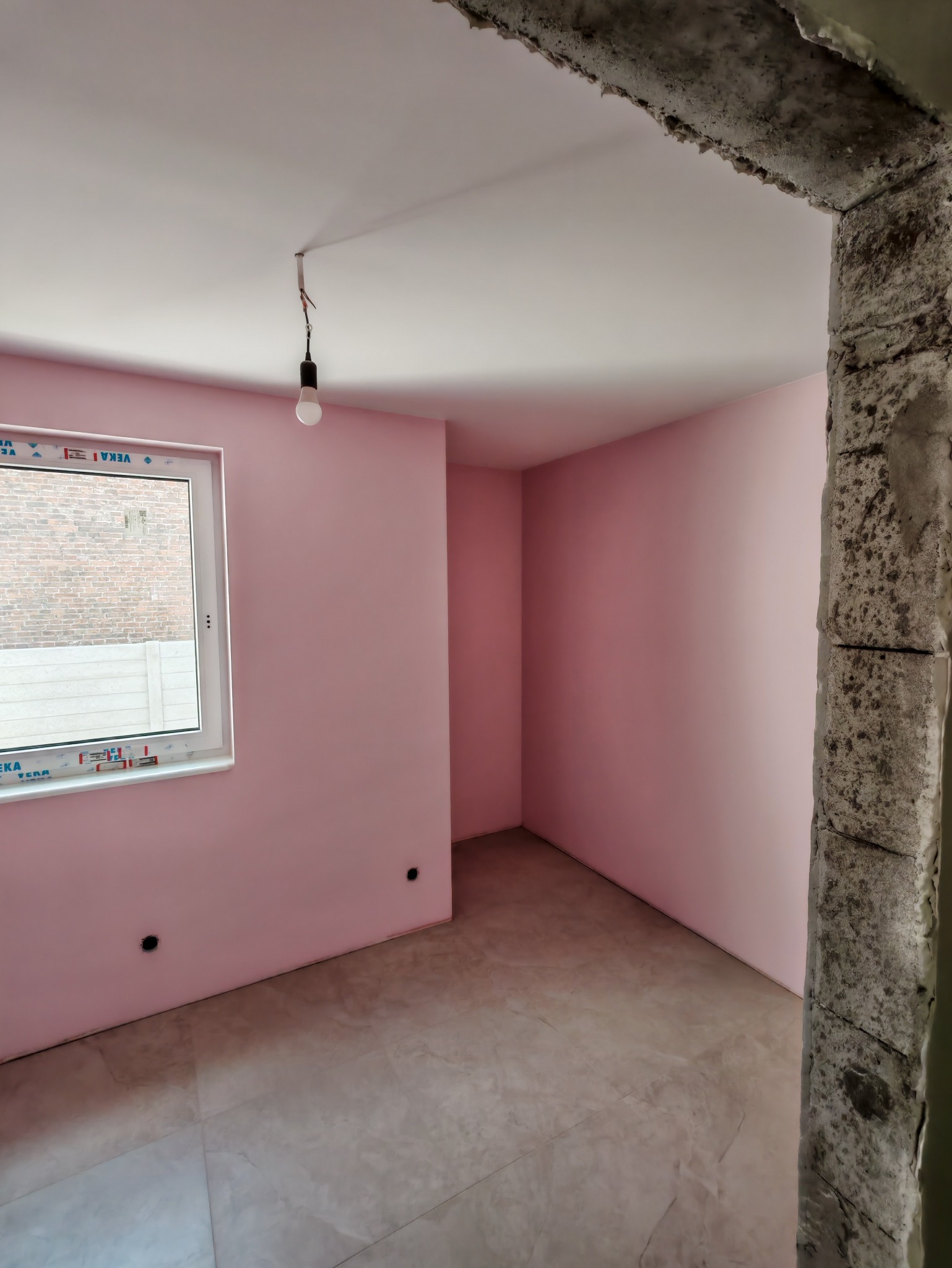Pokój po malowaniu ścian na różowo, z widocznym oknem i surowym wykończeniem framugi drzwi. Podłoga wyłożona płytkami. Wisząca żarówka.