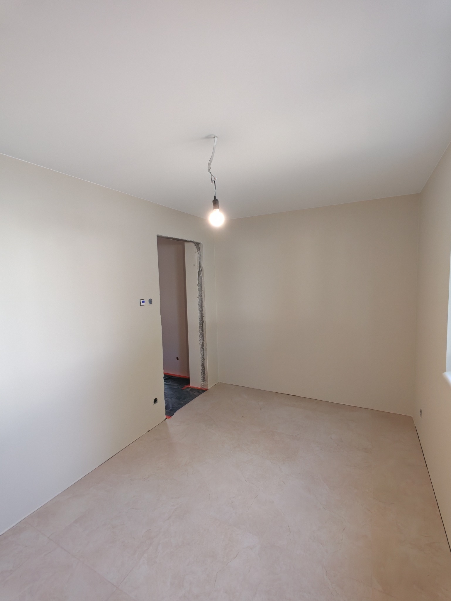 Jasne, świeżo pomalowane wnętrze z beżowymi ścianami i podłogą z płytek. Widoczne gniazdka elektryczne i surowa ościeżnica drzwiowa z widoczną instalacją elektryczną.