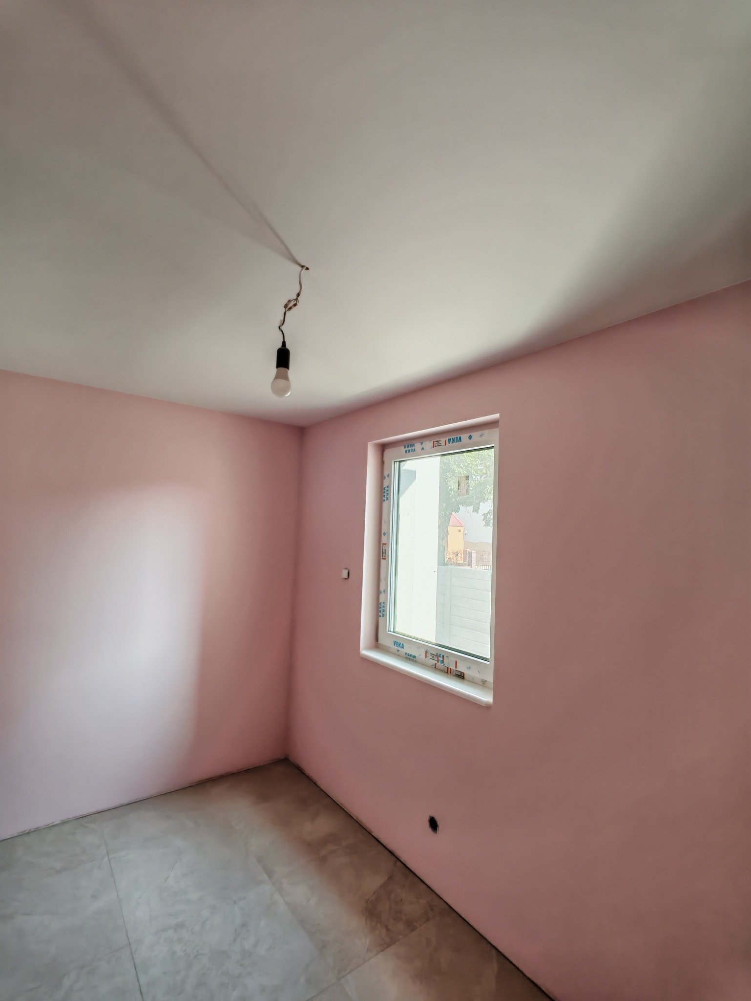 Pokój po malowaniu na różowo z nowym oknem, widoczna wisząca żarówka i szare płytki na podłodze. Świeżo pomalowane ściany i sufit.