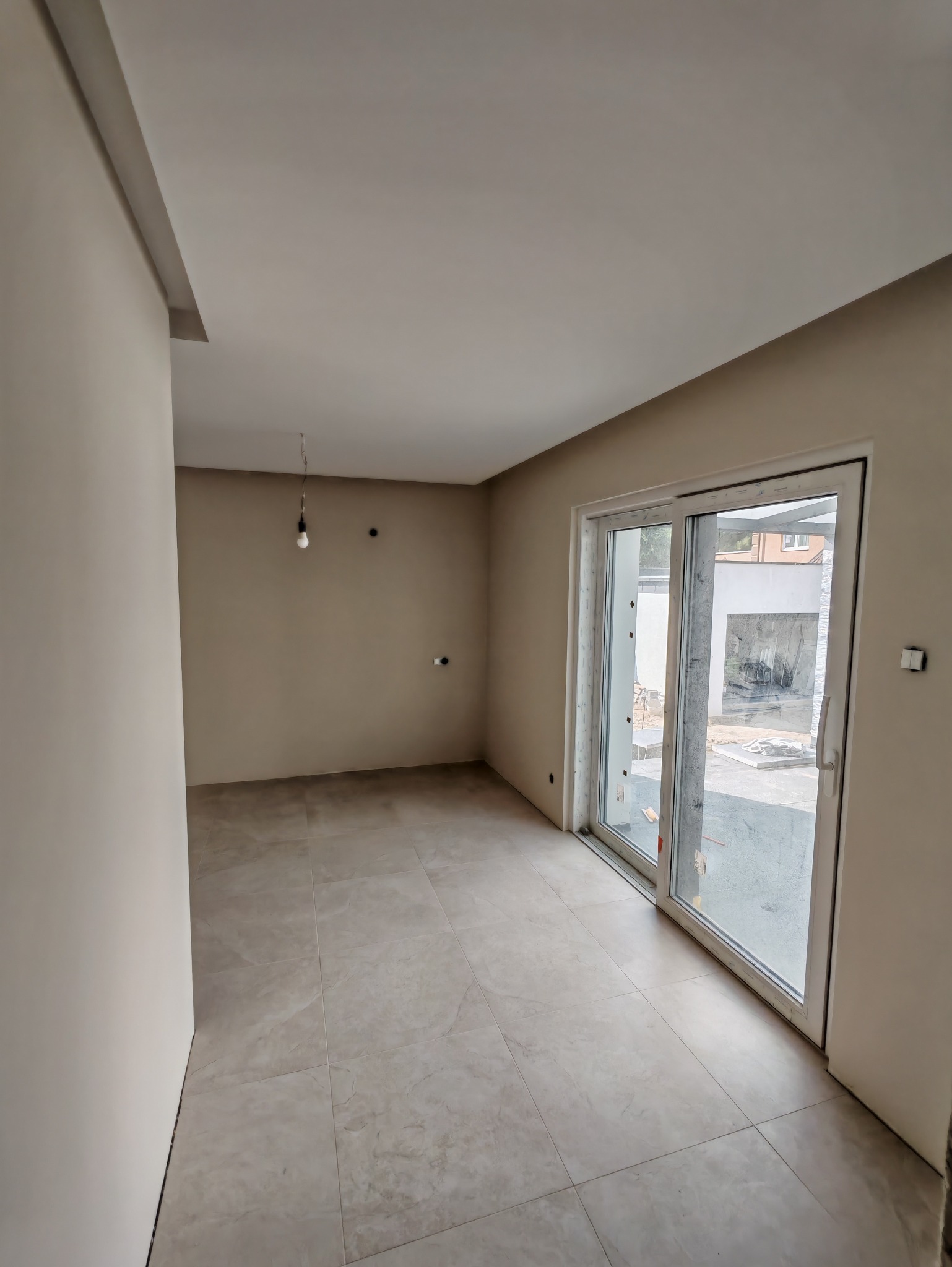 Jasne, beżowe wnętrze po remoncie: świeżo pomalowane ściany, płytki podłogowe imitujące marmur, duże przesuwne okno balkonowe w białej ramie.