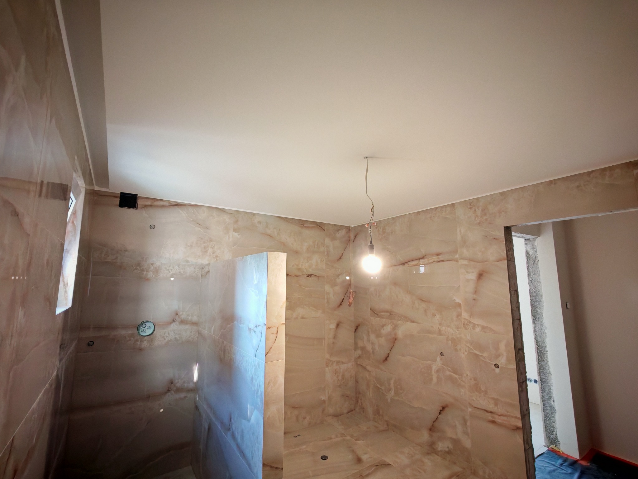 Wykończona łazienka z beżowymi płytkami imitującymi marmur, kabina prysznicowa w trakcie montażu, widoczne punkty świetlne i sufit podwieszany.