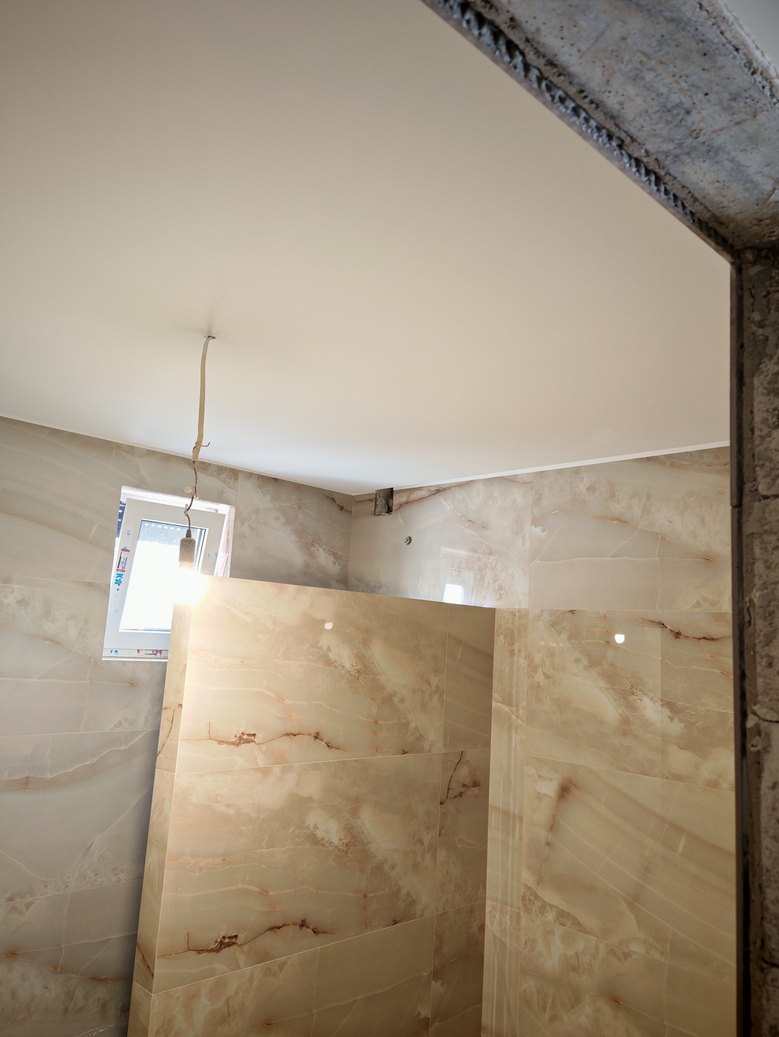 Wykończona łazienka z beżowymi płytkami imitującymi marmur i białym sufitem napinanym. Widoczne niedokończone wykończenie futryny drzwiowej.