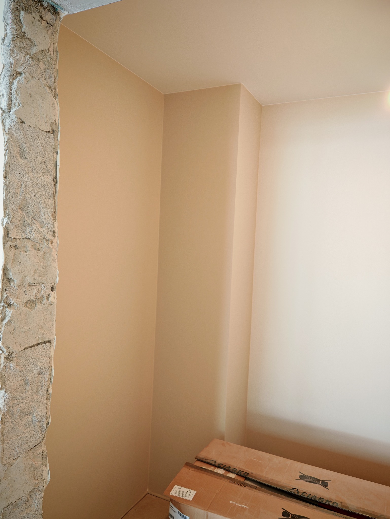 Wykończony róg pokoju po pracach w Gniezno: gładkie, beżowe ściany kontrastują z surowym betonem futryny. Na podłodze kartony z materiałami wykończeniowymi.