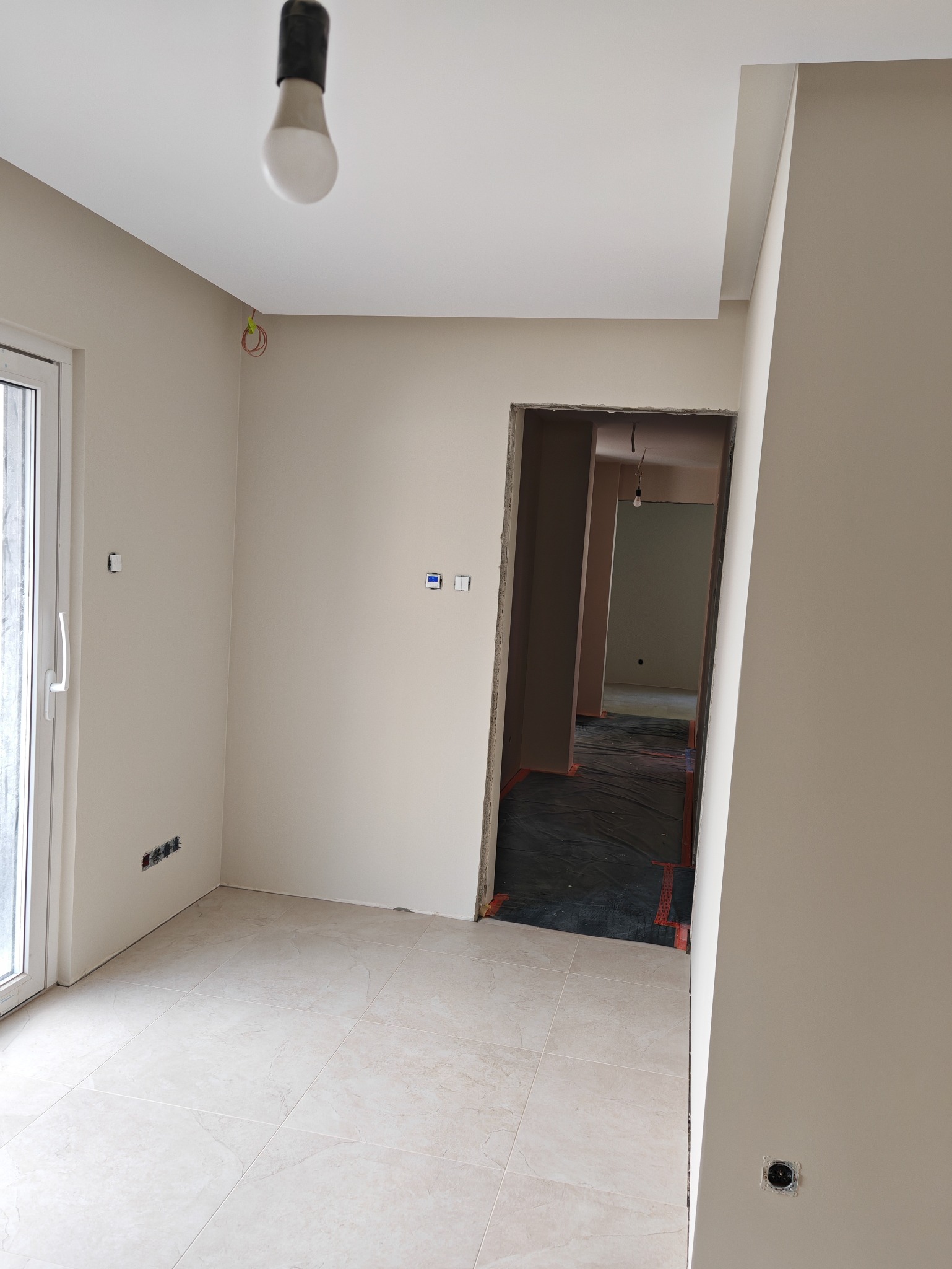 Jasne, kremowe ściany świeżo wykończonego pokoju z terakotą. Widoczne gniazdka i włączniki. W tle przejście do kolejnego pomieszczenia z zabezpieczoną podłogą.