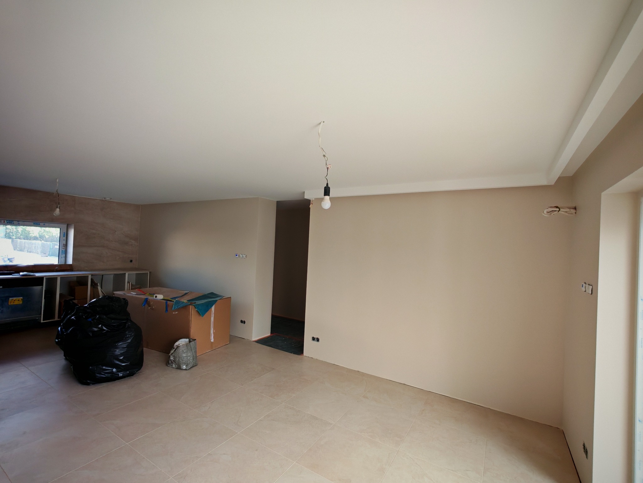 Jasne wnętrze po szpachlowaniu ścian i sufitu, widoczne gładkie powierzchnie i przygotowane podłoże pod wykończenie. Fragment okna, podłogi z płytek i prowizoryczne oświetlenie.