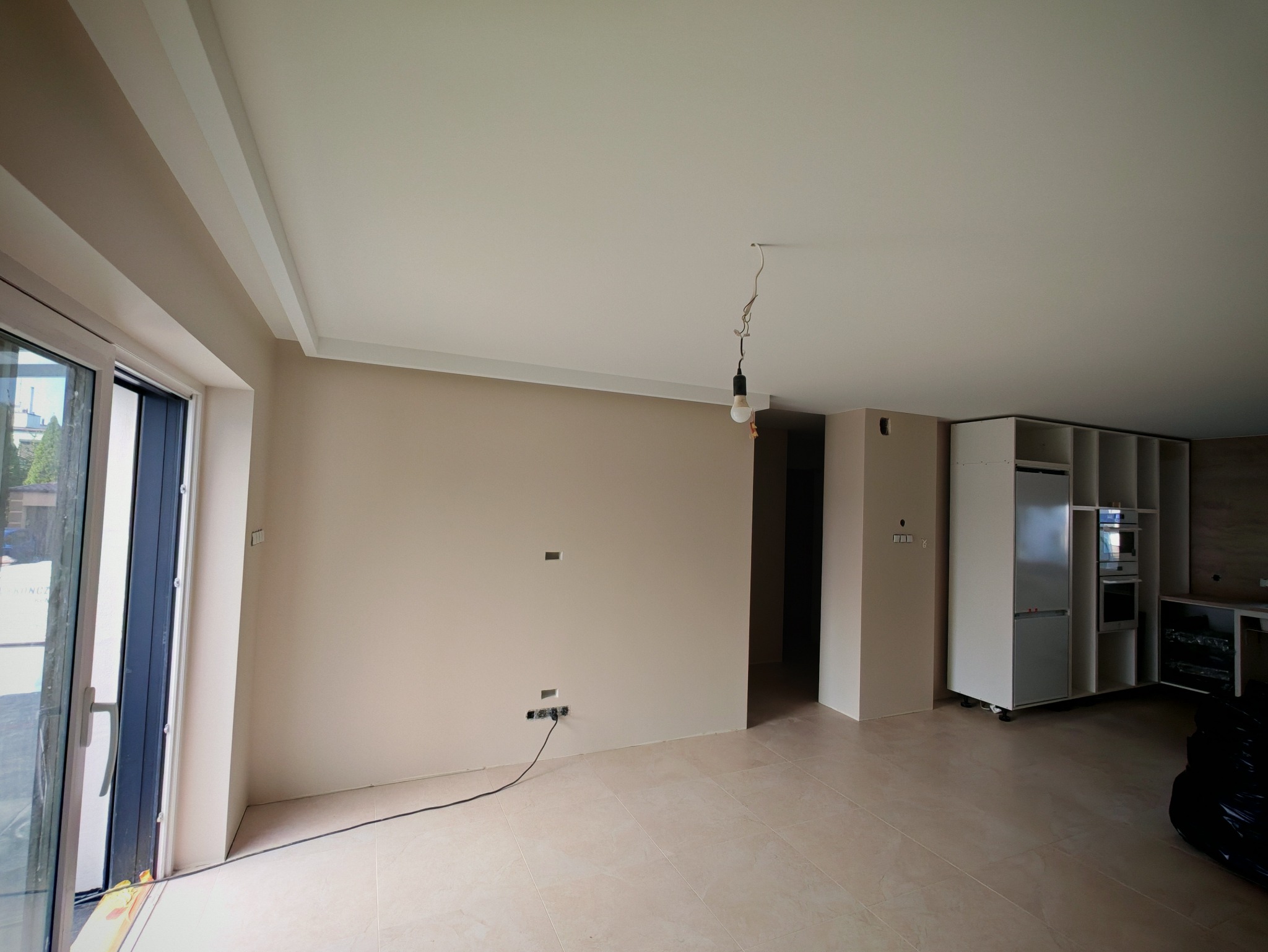 Jasne wnętrze w trakcie wykańczania: beżowe ściany, biały sufit z wystającą żarówką, częściowo zabudowana biała kuchnia. Widoczne gniazdka elektryczne.