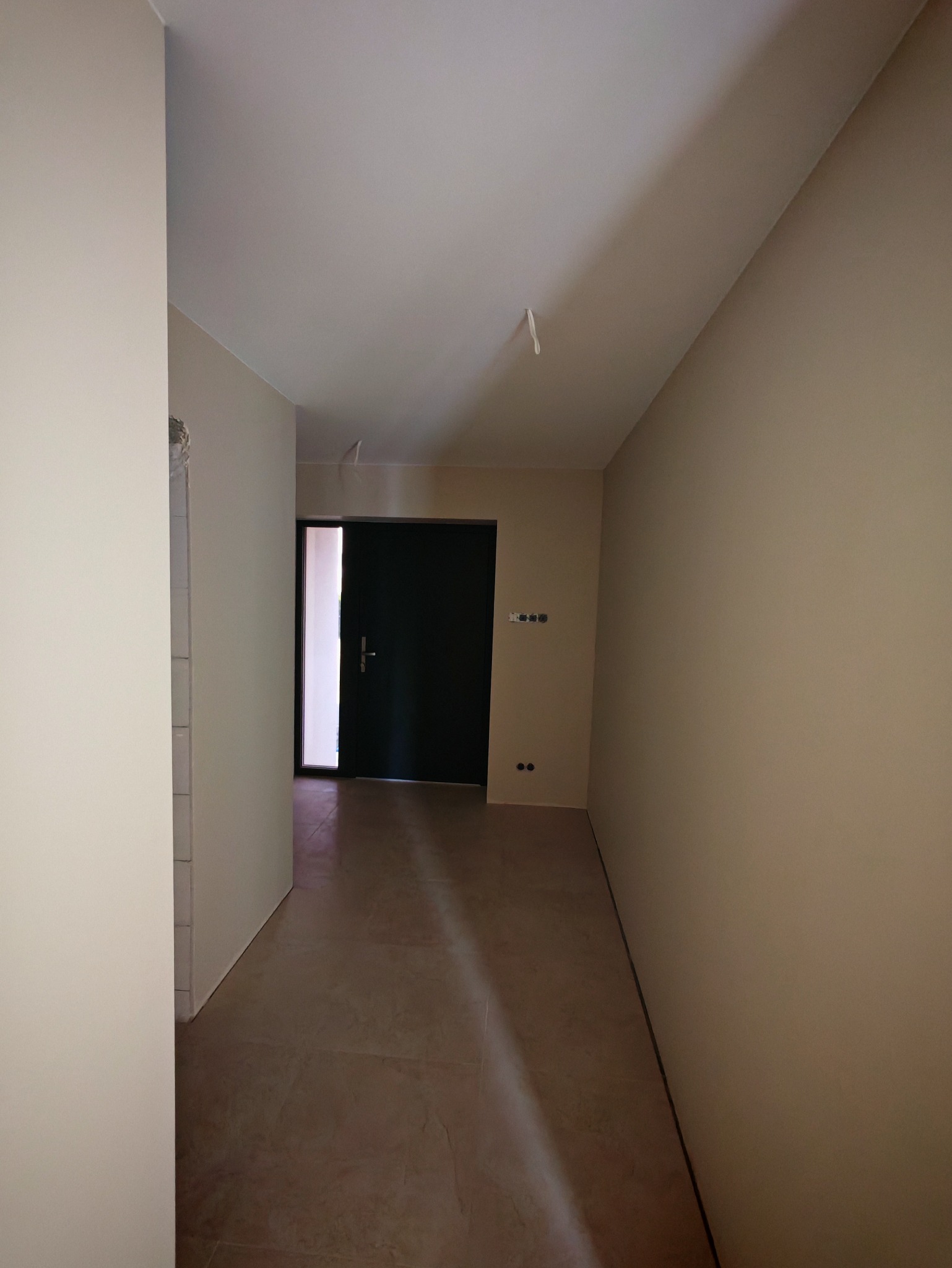 Korytarz po remoncie: beżowe ściany, ciemne drzwi, brązowa podłoga. Widoczne gniazdka i kable wystające z sufitu. Minimalistyczny design z surowym wykończeniem.