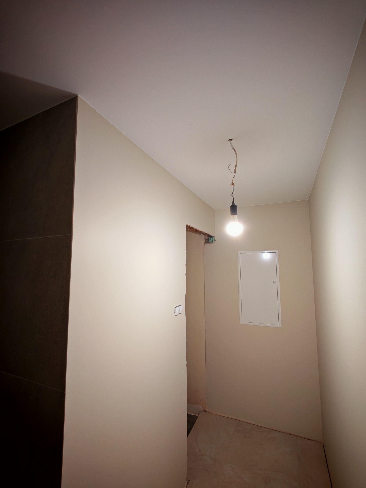 Korytarz po malowaniu ścian na delikatny beż, widoczna instalacja elektryczna i otwór drzwiowy w trakcie wykańczania. Na suficie zwisa żarówka.