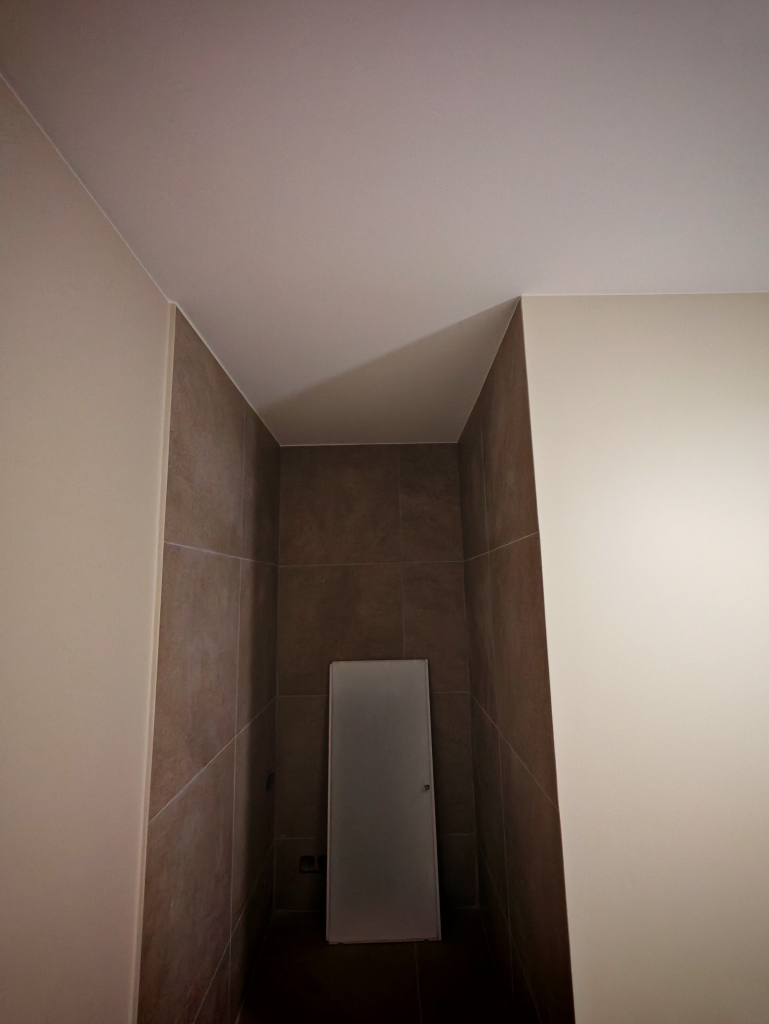 Wnęka wyłożona brązowymi płytkami, z małą, szarą szafką w tle. Ściany po bokach w odcieniach beżu, sufit biały. Minimalistyczne wykończenie łazienki.