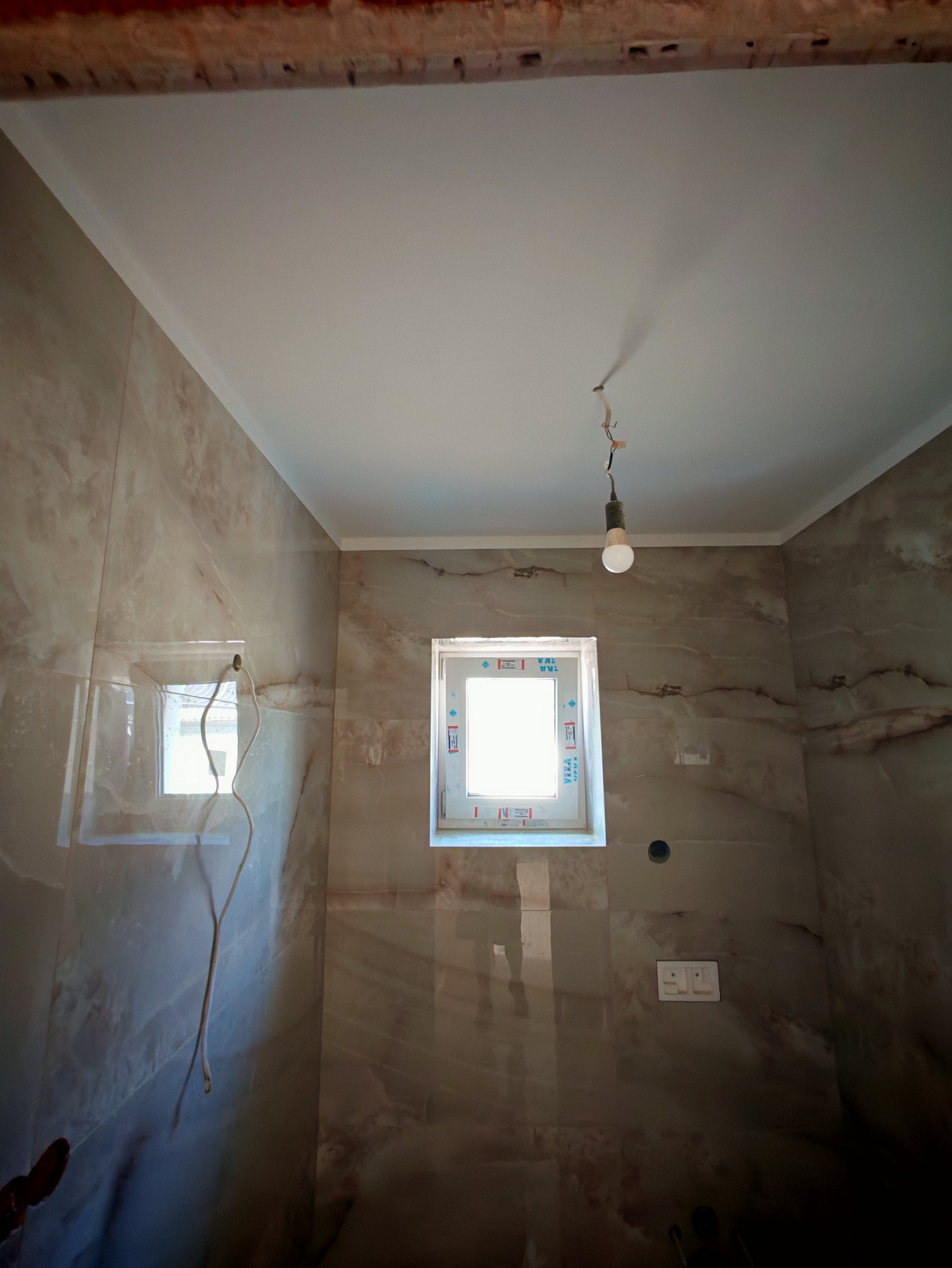 Wykończona łazienka z marmurowymi płytkami, białym sufitem i oknami. Widoczna żarówka na kablu. Prace wykończeniowe.