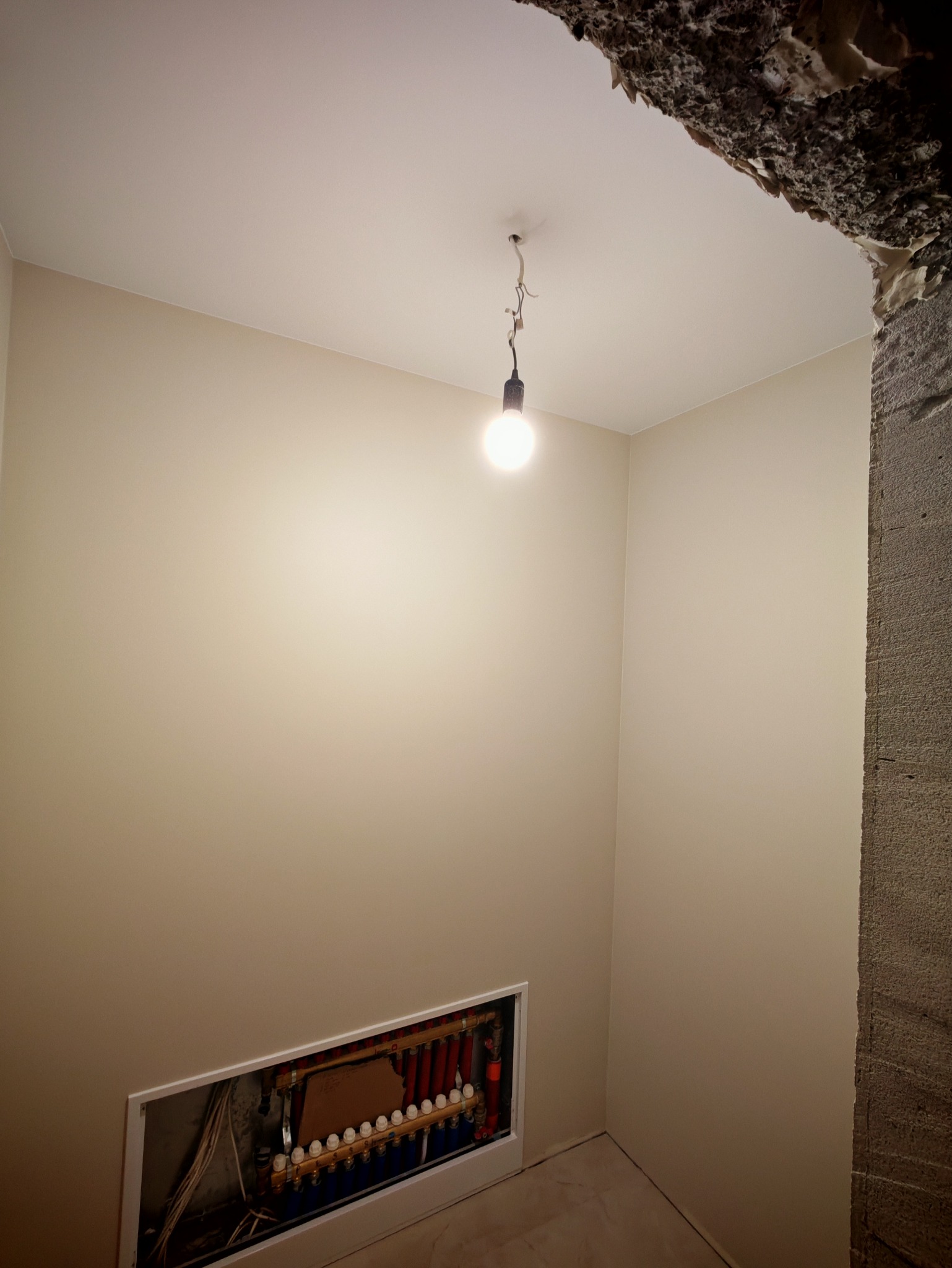 Wnętrze po remoncie: gładkie, beżowe ściany, biały sufit z żarówką na kablu i ukryta instalacja ogrzewania podłogowego. Fragment surowego betonu w rogu.