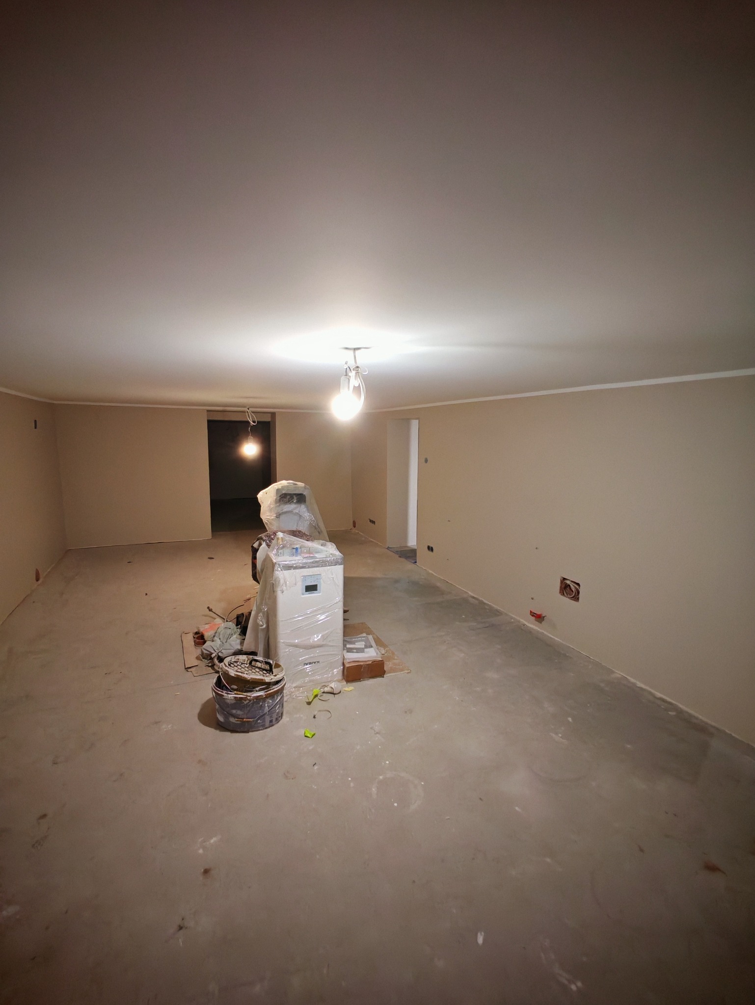 Beżowe ściany w pustym pokoju po szpachlowaniu. Na podłodze wiadro i maszyna w folii ochronnej. Widoczne gniazdka elektryczne.