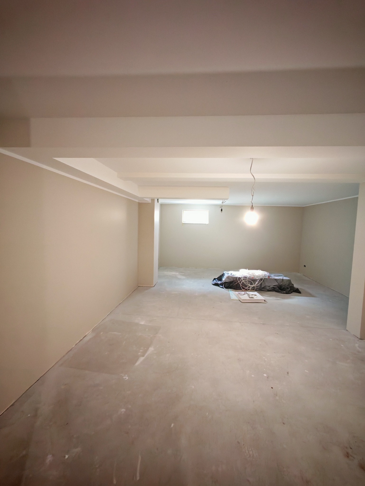 Jasne wnętrze w trakcie odświeżania: ściany pomalowane na beżowo, sufit biały z widocznymi belkami, podłoga surowa. Kabel oświetleniowy zwisa z sufitu.