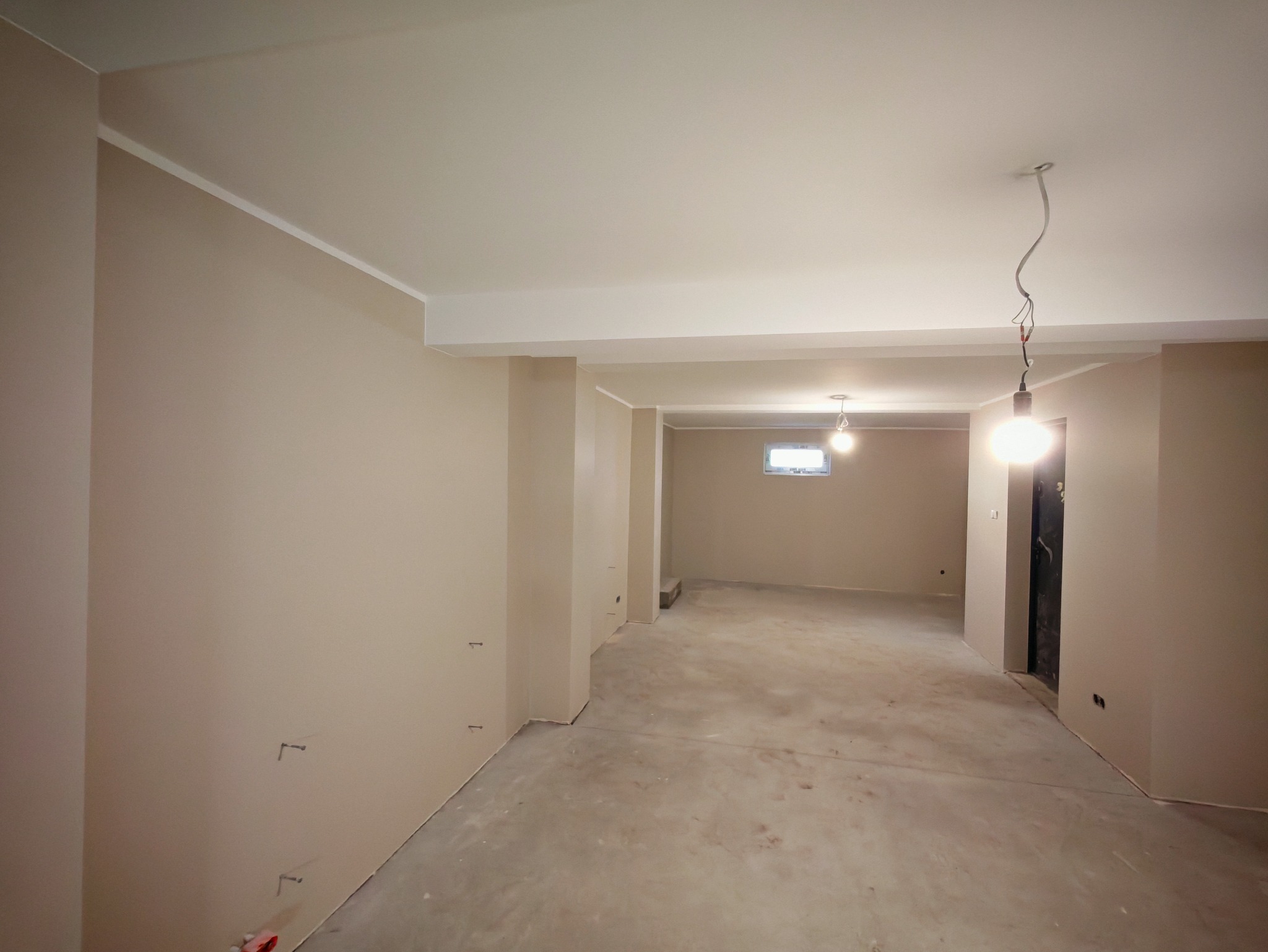 Wnętrze po szpachlowaniu i malowaniu na beżowo, widoczne listwy przypodłogowe i sufitowe. Na suficie wiszące żarówki. Betonowa podłoga przed wykończeniem.