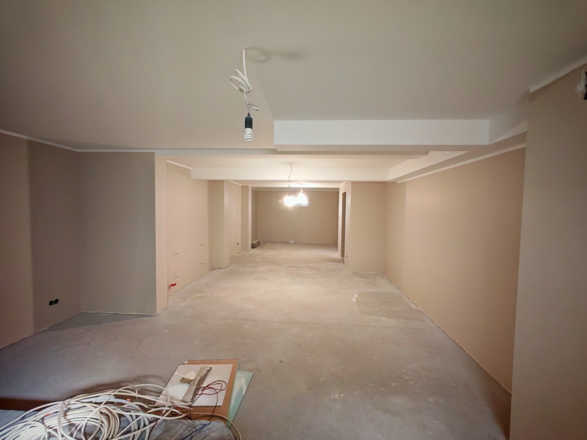 Wnętrze w trakcie wykończenia: świeżo pomalowane ściany w beżowym odcieniu, surowa podłoga, widoczne przewody i narzędzia. Oświetlenie tymczasowe.