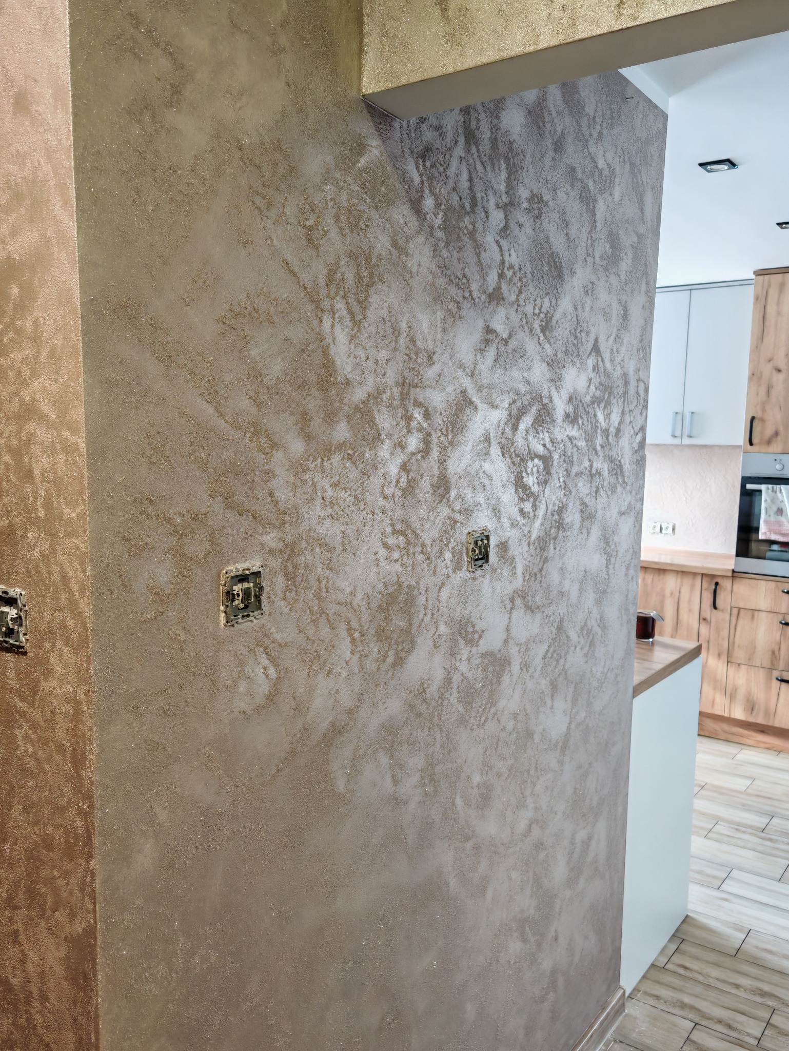 Dekoracyjna ściana w odcieniach beżu i bieli z efektem marmuru, widoczne gniazdka elektryczne. W tle fragment kuchni z drewnianymi szafkami.