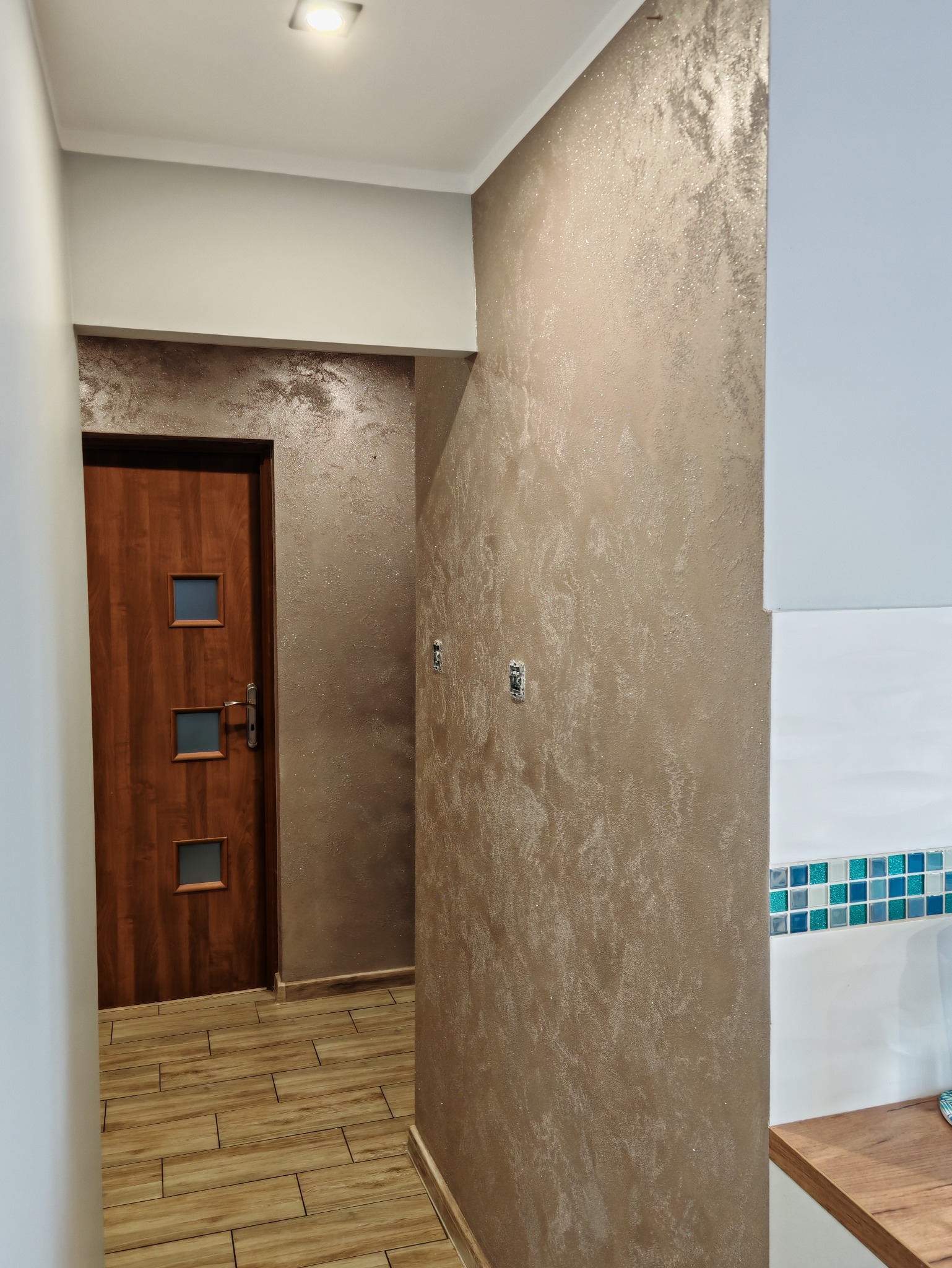 Efektowna, dekoracyjna ściana w odcieniach beżu z połyskiem w korytarzu w Gnieznie. Widoczna drewniana podłoga i drzwi z szybkami.