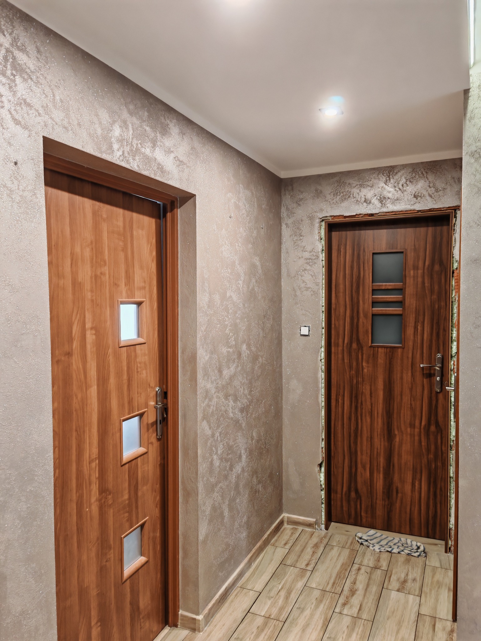 Korytarz po remoncie z dwoma drewnianymi drzwiami, ściany pokryte dekoracyjnym tynkiem w odcieniach beżu i szarości, podłoga wyłożona płytkami imitującymi drewno.