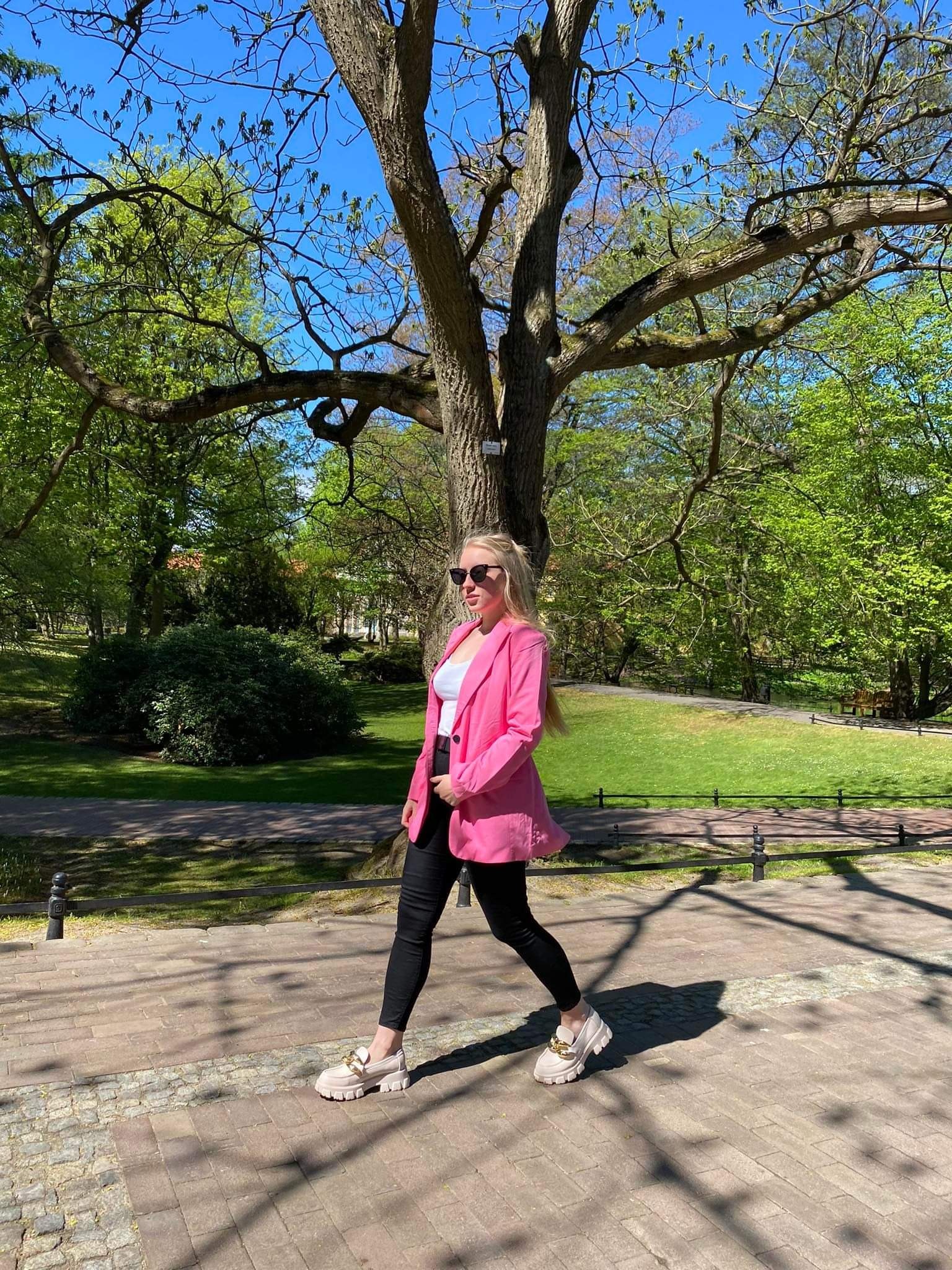 Blondwłosa kobieta w różowej marynarce i czarnych spodniach spaceruje po brukowanym chodniku w parku, na tle drzew i zieleni.