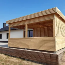 Nowoczesna, drewniana konstrukcja z poziomych desek, pełniąca funkcję zadaszenia tarasu, z widocznymi elementami montażowymi i fragmentem domu w tle, na tle błękitnego nieba.