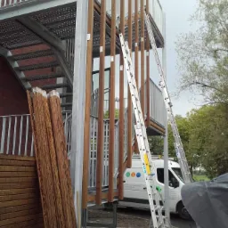 Konstrukcja schodów zewnętrznych z elementami drewnianymi i metalowymi w trakcie montażu, widoczne drabiny, zaparkowany biały samochód dostawczy w tle.