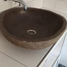 Umywalka z naturalnego kamienia w łazience z białymi płytkami i mozaiką na ramie lustra. Bateria chromowana, widoczny fragment podłogi z szarych płytek.