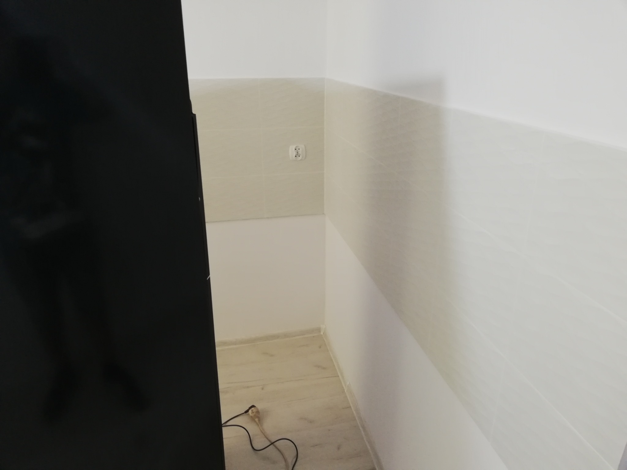 Wykończone wnętrze pomieszczenia z beżowymi płytkami do połowy ściany, widoczne gniazdko elektryczne, fragment czarnego mebla i kabel na podłodze z jasnymi panelami.