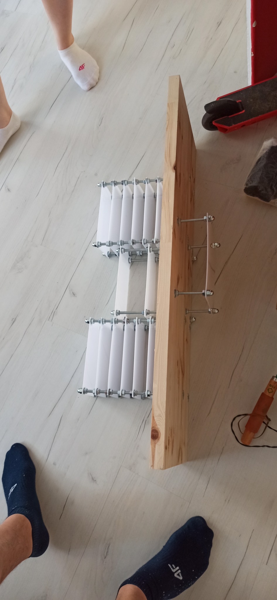 Element konstrukcyjny, prawdopodobnie fragment schodów strychowych, z drewnianą belką i białymi, pionowymi płytkami przymocowanymi śrubami, widoczne stopy w skarpetkach na podłodze imitującej drewno.