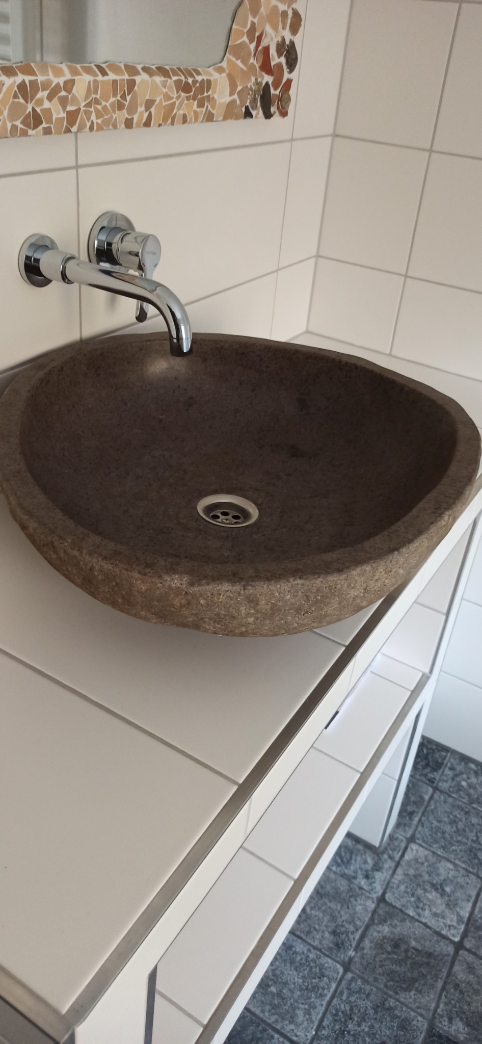 Umywalka z naturalnego kamienia w łazience z białymi płytkami i mozaiką na ramie lustra. Bateria chromowana, widoczny fragment podłogi z szarych płytek.