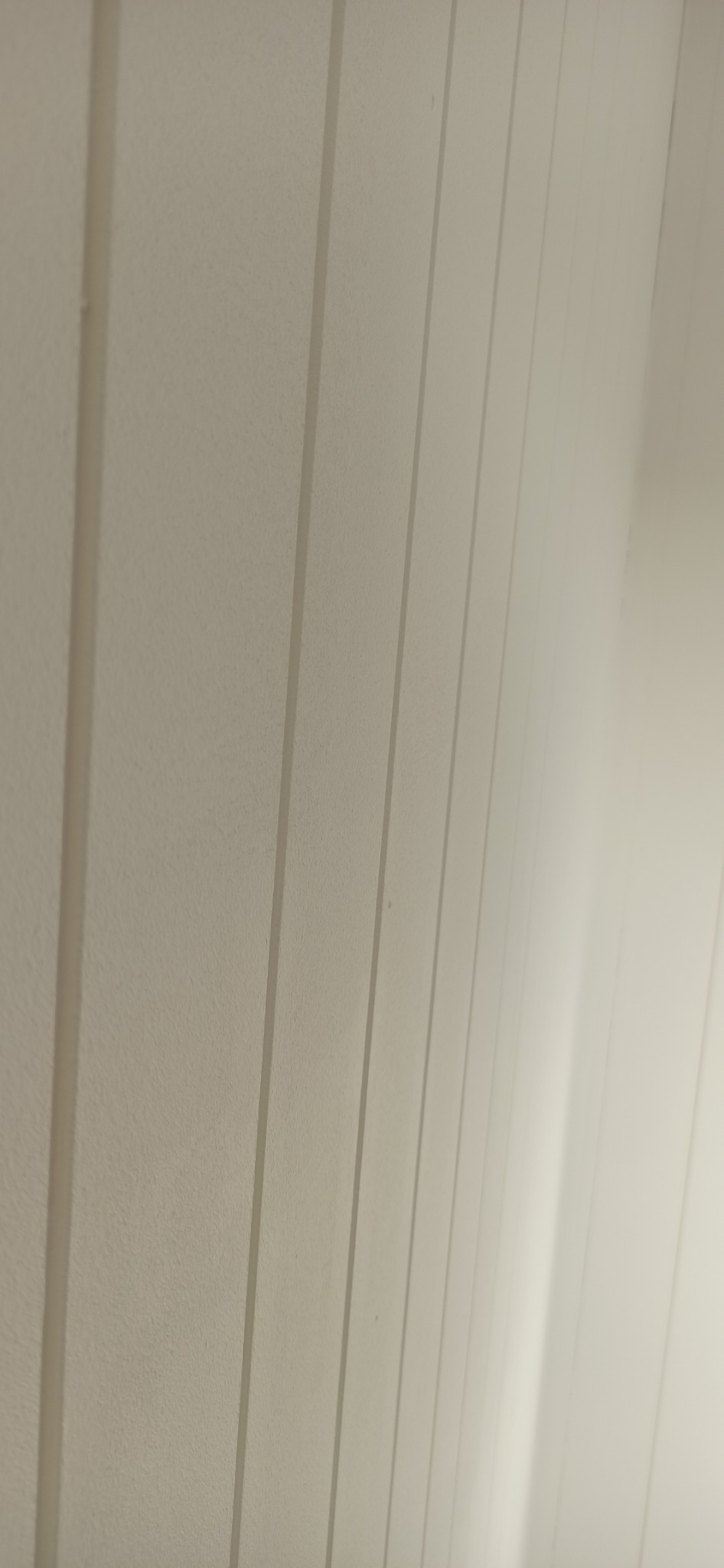 Ściana z pionowymi panelami pomalowana na jasny, neutralny kolor, widoczna pod kątem, z delikatną fakturą i subtelnym cieniowaniem.