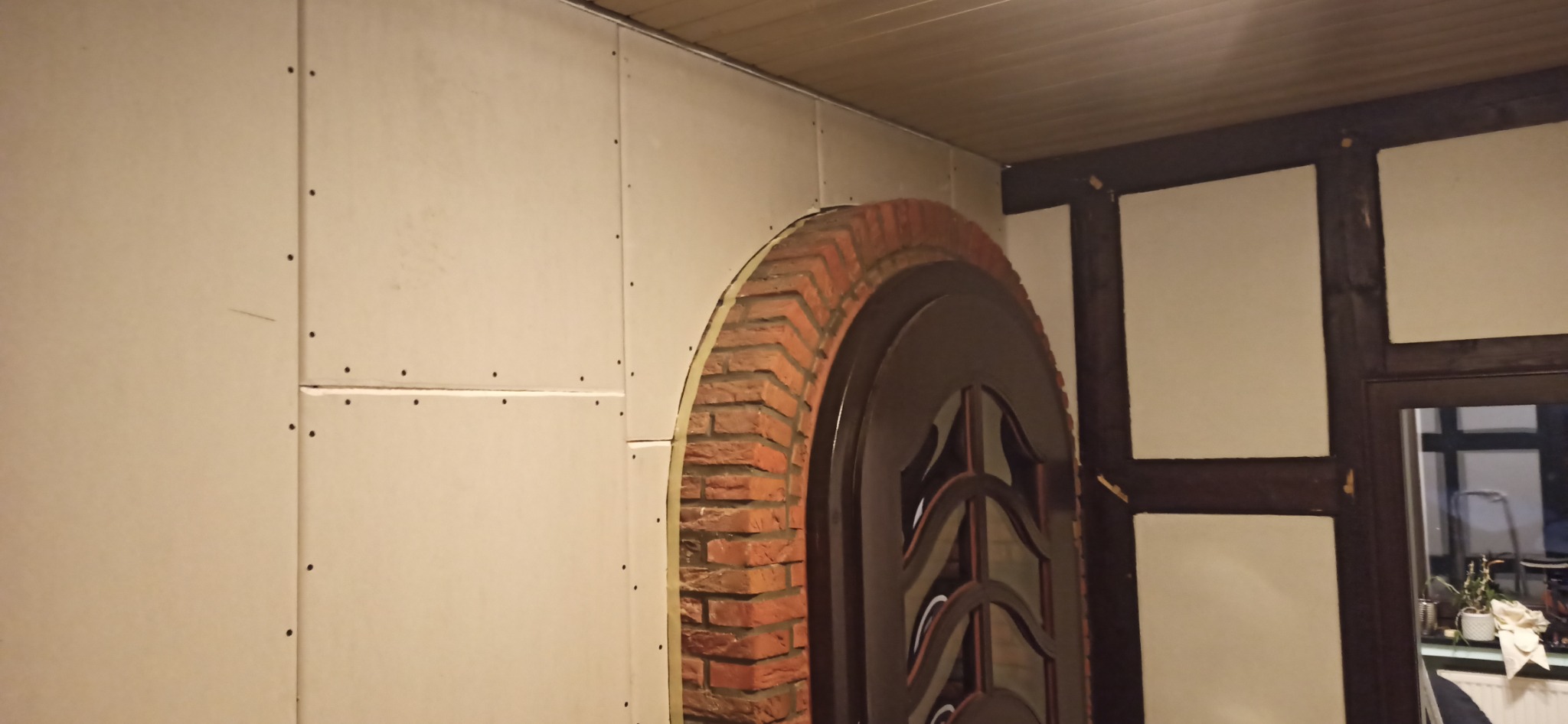 Wnętrze w trakcie prac: zamontowane płyty gipsowo-kartonowe z widocznymi łączeniami i śrubami, obok łukowatego otworu obłożonego cegłą, w tle fragment konstrukcji szkieletowej ściany.