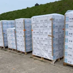 Siedem palet z zapakowanymi kartonami UNICARE, ustawionych w rzędzie na betonowym placu, z wózkiem paletowym i samochodem BMW w tle.