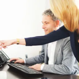 Mężczyzna w szarej marynarce pracuje przy komputerze, kobieta w granatowej marynarce wskazuje palcem na ekran monitora. Na biurku telefon stacjonarny, klawiatura, myszka oraz miniaturowe figurki.