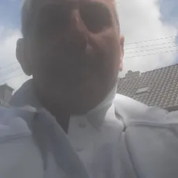 Autoportret mężczyzny w białej koszuli polo na tle nieba i dachu domu.