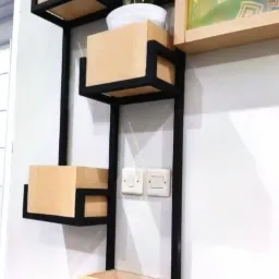 Półka ścienna z czterema drewnianymi kubikami w czarnej, metalowej ramie, na drugim od góry kubiku roślina w białej doniczce, obok rama z obrazem, na ścianie widoczne włączniki światła.