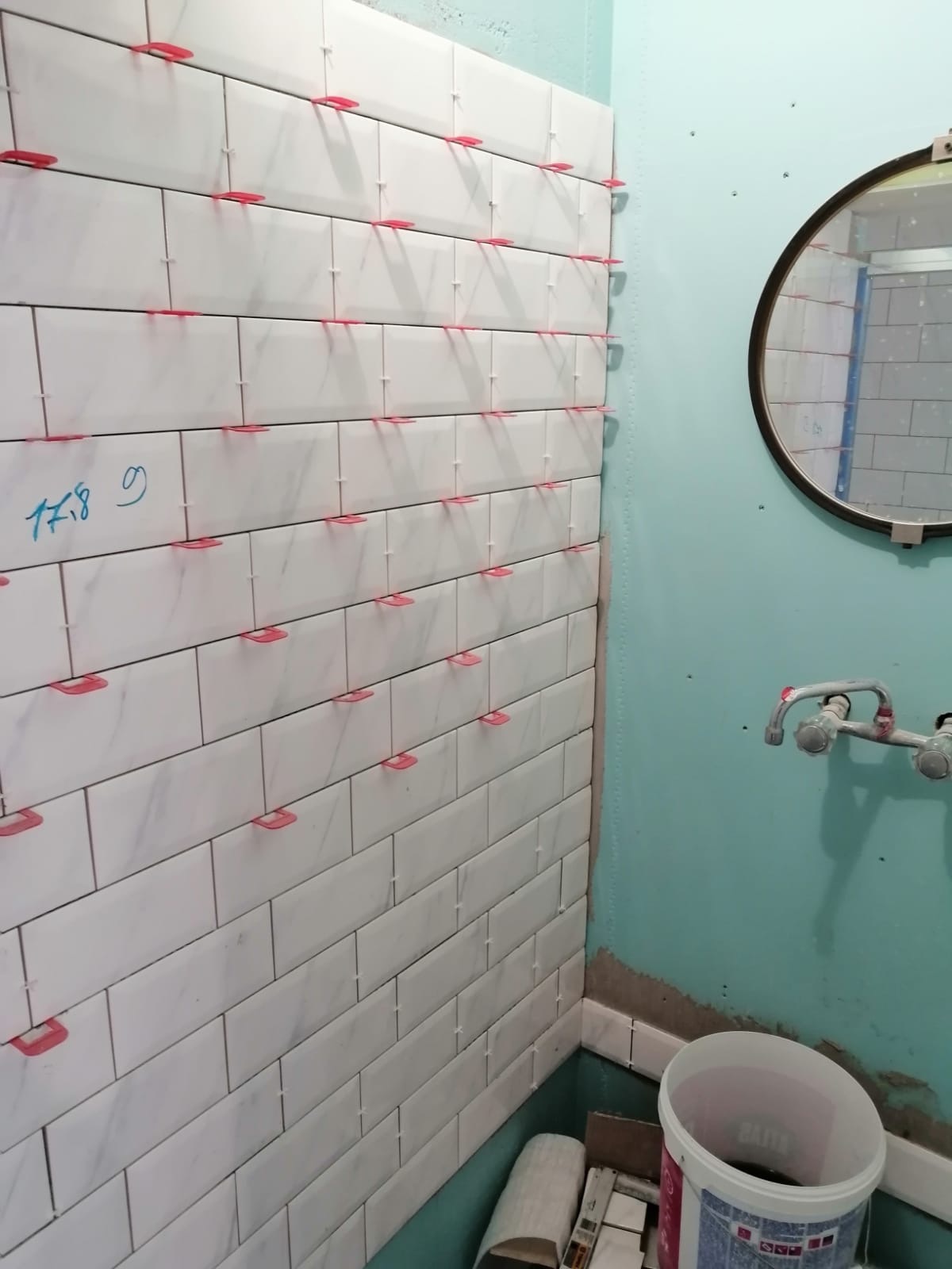 Świeżo ułożone białe, marmurkowe płytki w kształcie cegiełek na ścianie łazienki z systemem poziomowania płytek; widoczne niebieskie ściany, okrągłe lustro i bateria umywalkowa