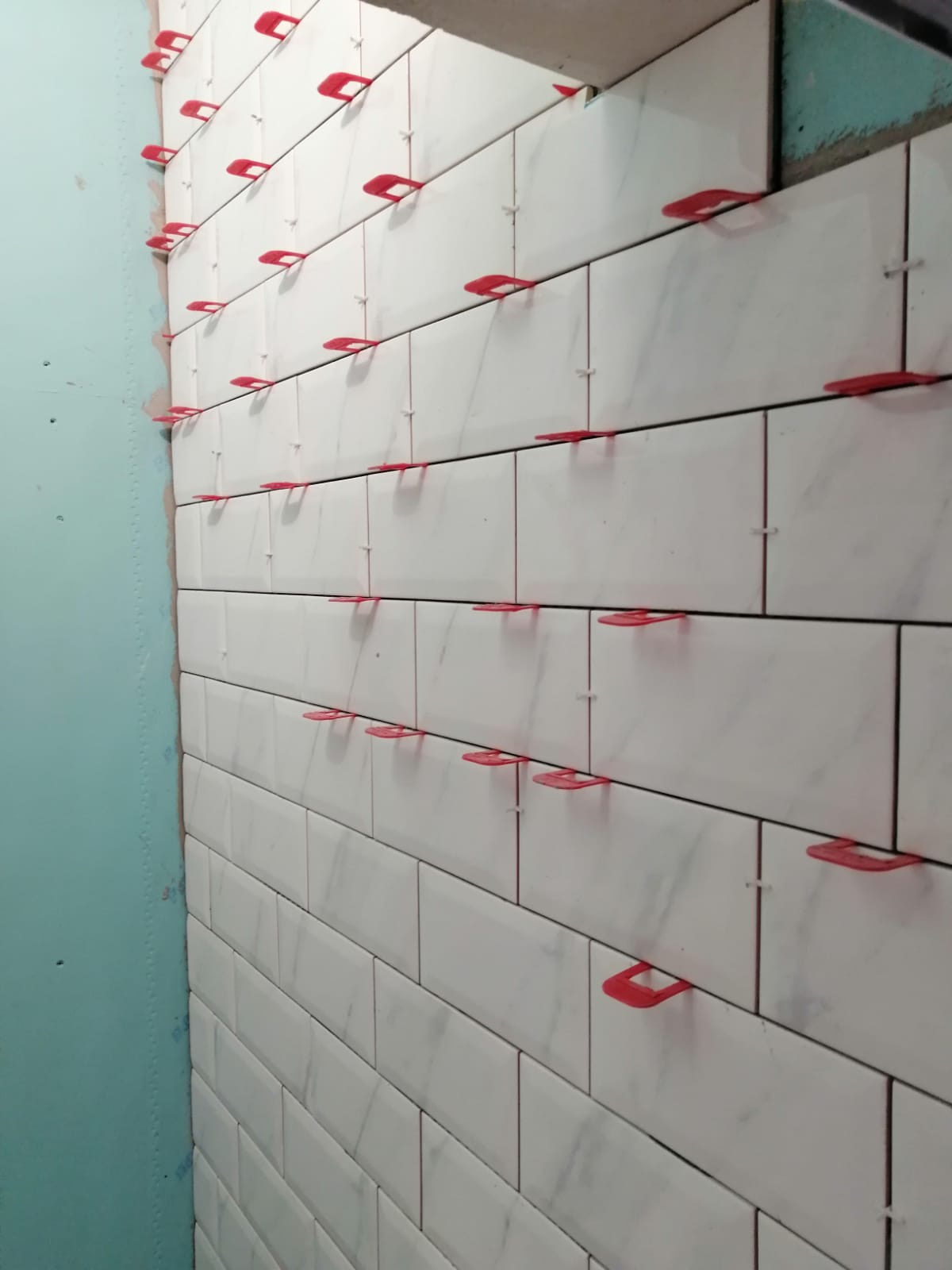 Ściana wyłożona białymi, podłużnymi płytkami imitującymi marmur, z czerwonymi klipsami poziomującymi spoiny, w trakcie prac glazurniczych.