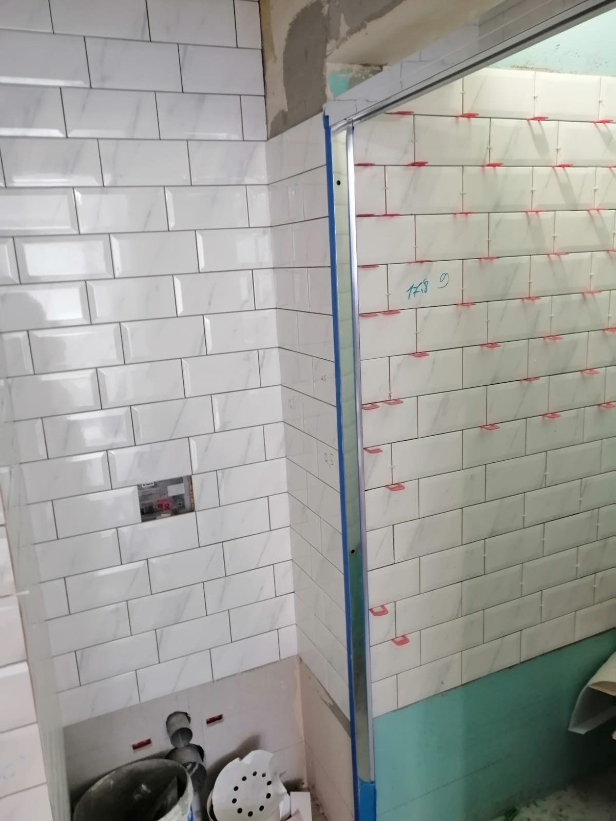 Białe, prostokątne płytki imitujące marmur, ułożone na ścianie łazienki, z widocznymi czerwonymi krzyżykami dystansowymi i zapisaną datą na jednej ze ścian, ukazujące etap prac wykończeniowych.