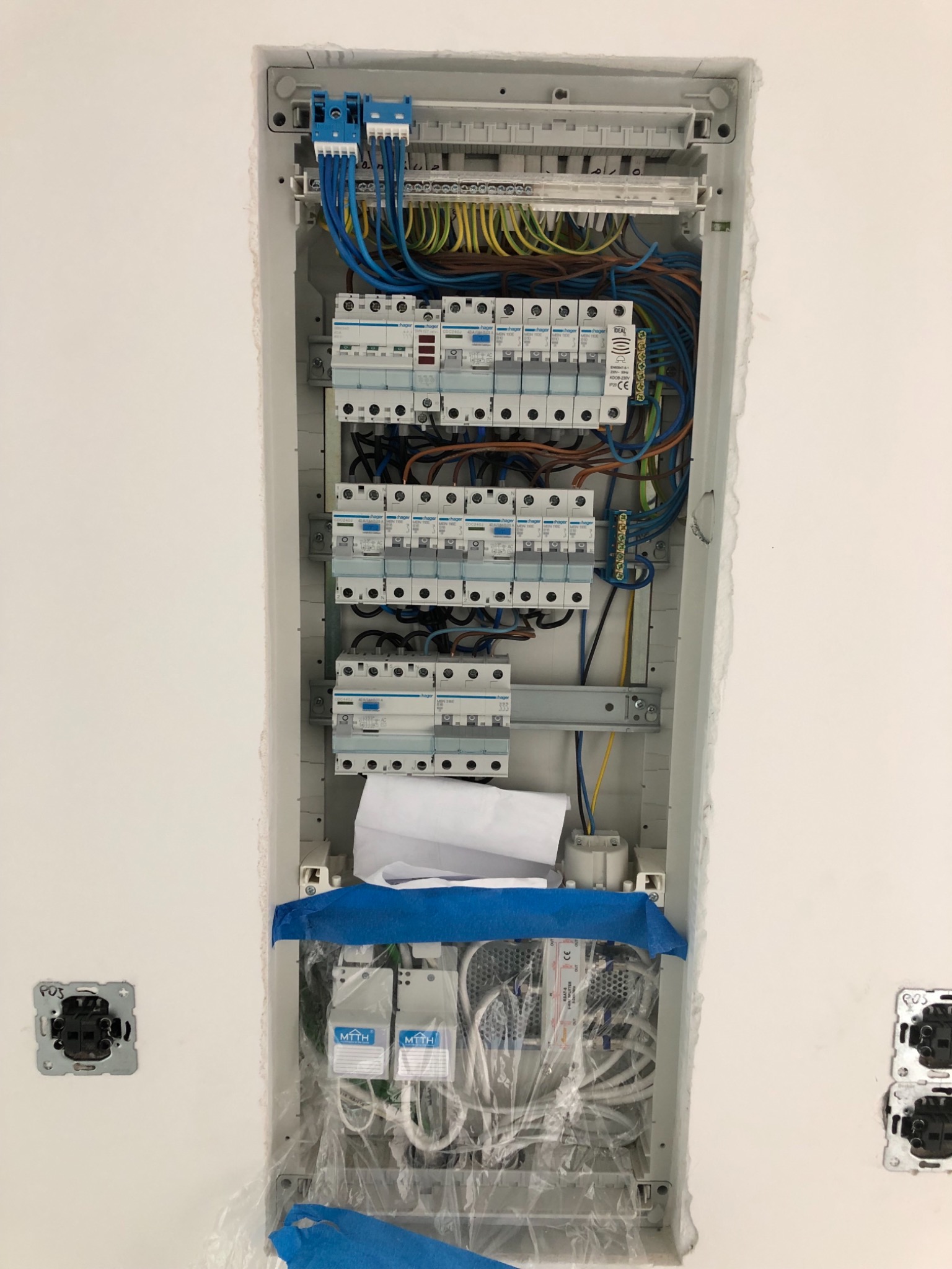 Rozdzielnia elektryczna w trakcie montażu w nowym budynku, widoczne zabezpieczenia folią i taśmą malarską, obok puszki elektryczne bez osprzętu.