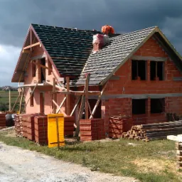 Surowy stan domu jednorodzinnego z czerwonej cegły z drewnianą konstrukcją dachu i stertami dachówek na zewnątrz w pochmurny dzień.