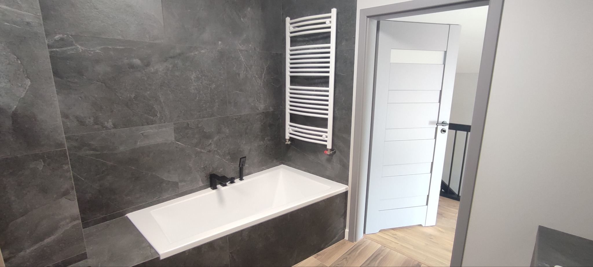 Nowoczesna łazienka z szarymi płytkami imitującymi kamień, białą wanną z czarną armaturą i białym grzejnikiem drabinkowym, widok na otwarte drzwi i drewnopodobną podłogę
