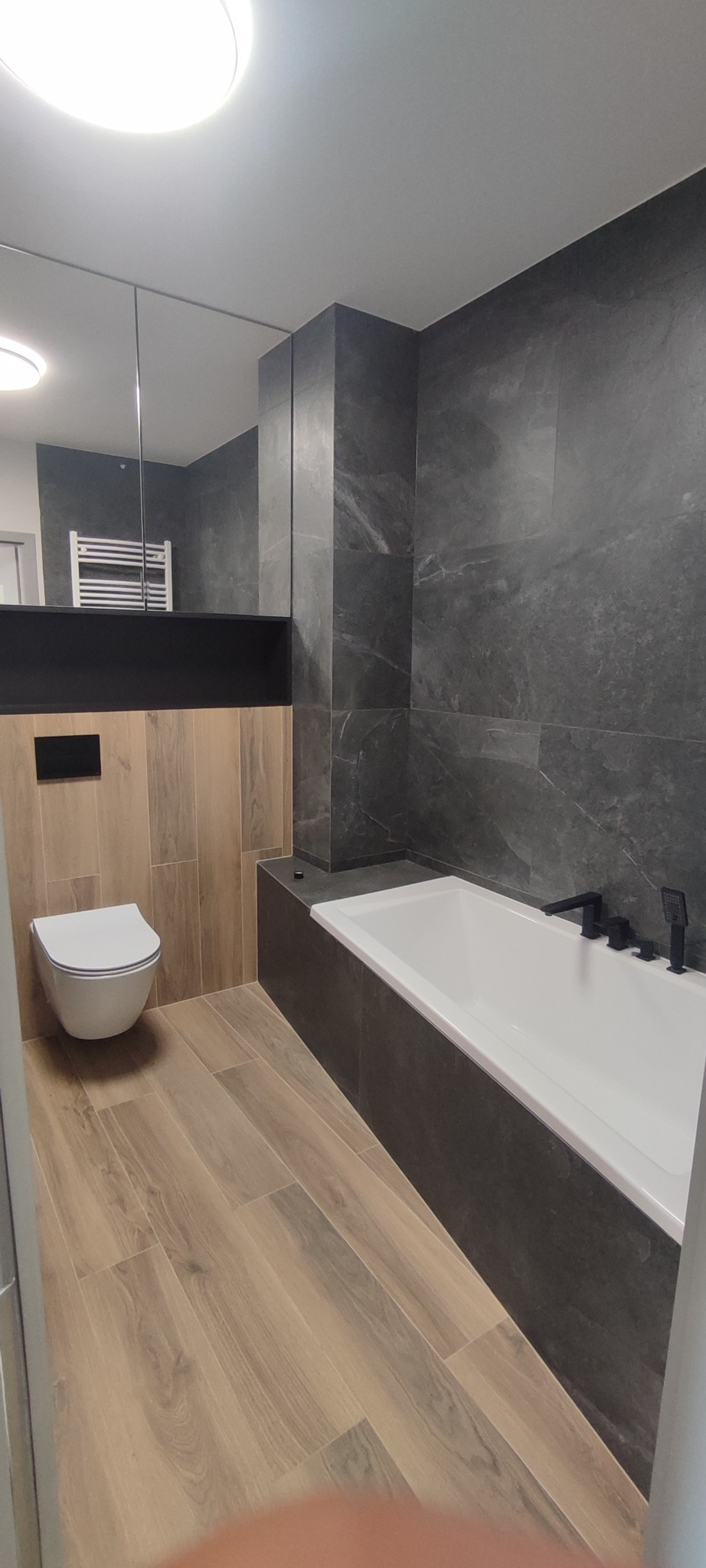 Nowoczesna aranżacja łazienki w Szczecinie: biała wanna z czarną armaturą, podwieszana toaleta, szare płytki imitujące kamień i drewnopodobna podłoga.