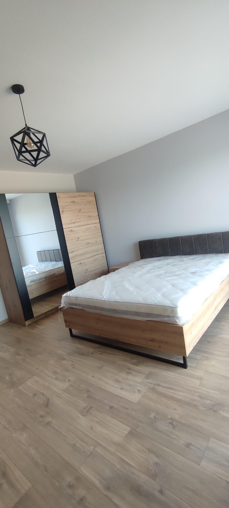 Nowoczesna sypialnia z drewnianą podłogą, łóżkiem z szarym zagłówkiem i szafą z lustrzanymi drzwiami, oświetlona geometryczną lampą wiszącą.